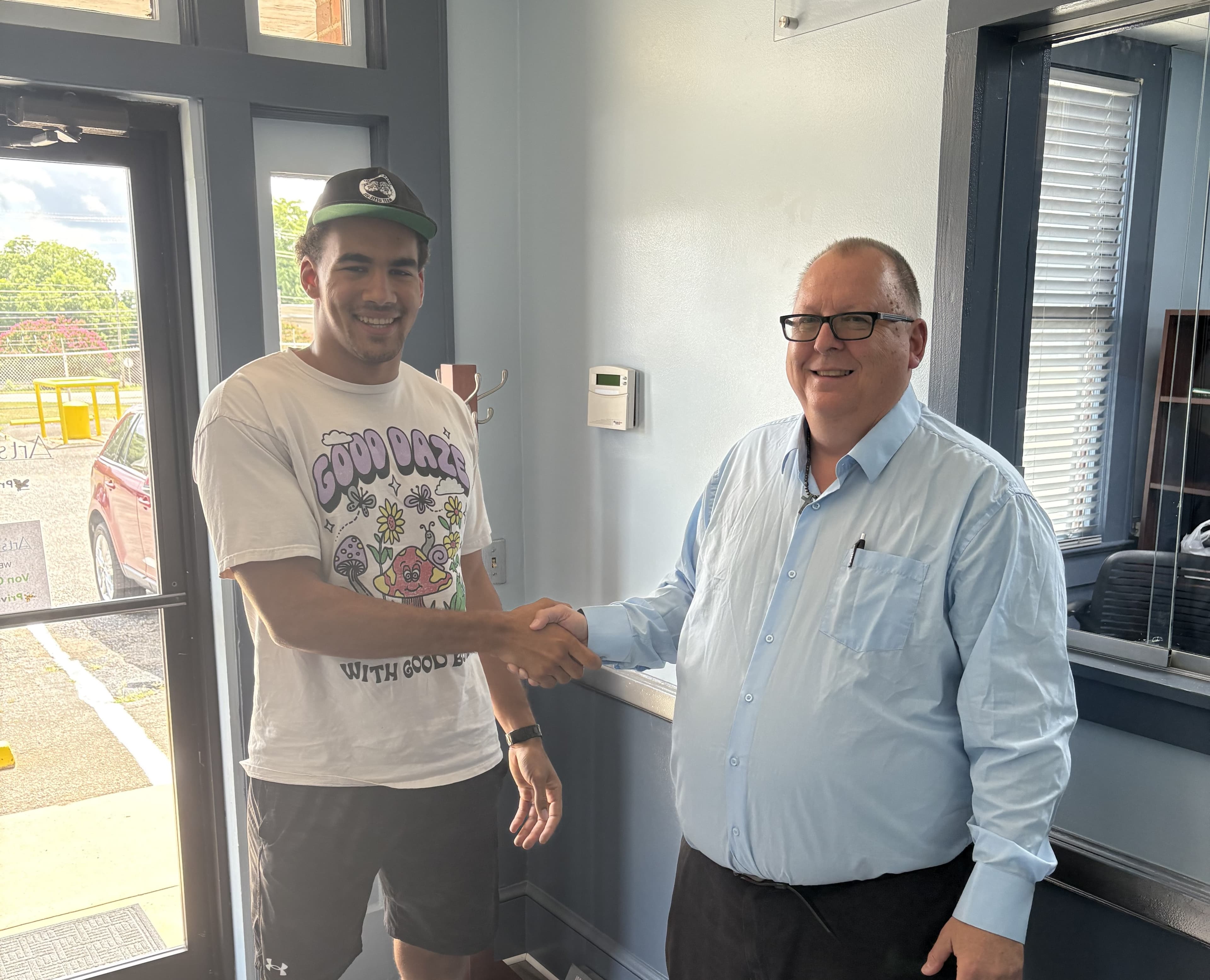 Smiling young man in a graphic t-shirt shakes hands with an older man in glasses.