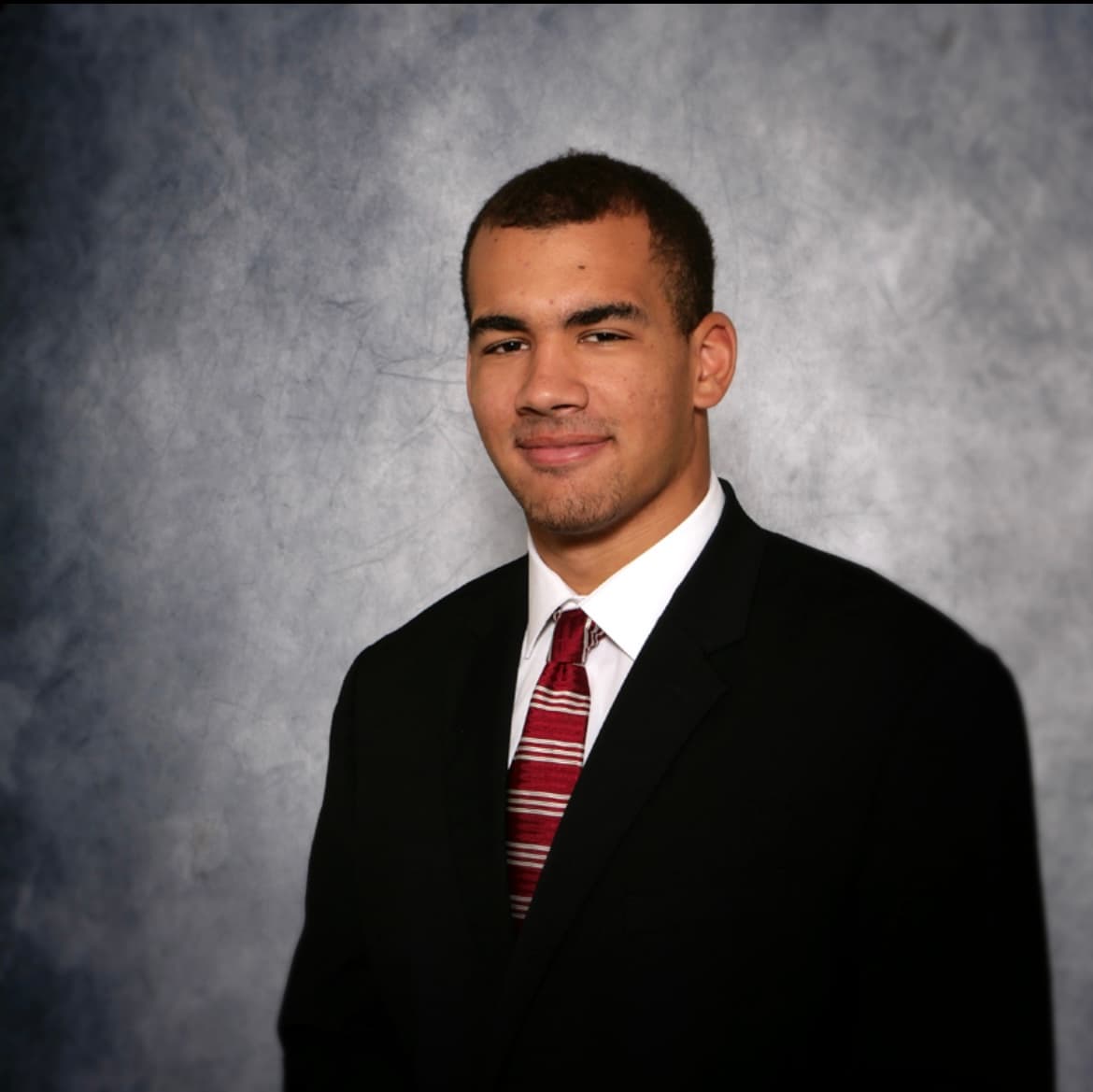 Young man in a black suit and red striped tie against a textured grey background.