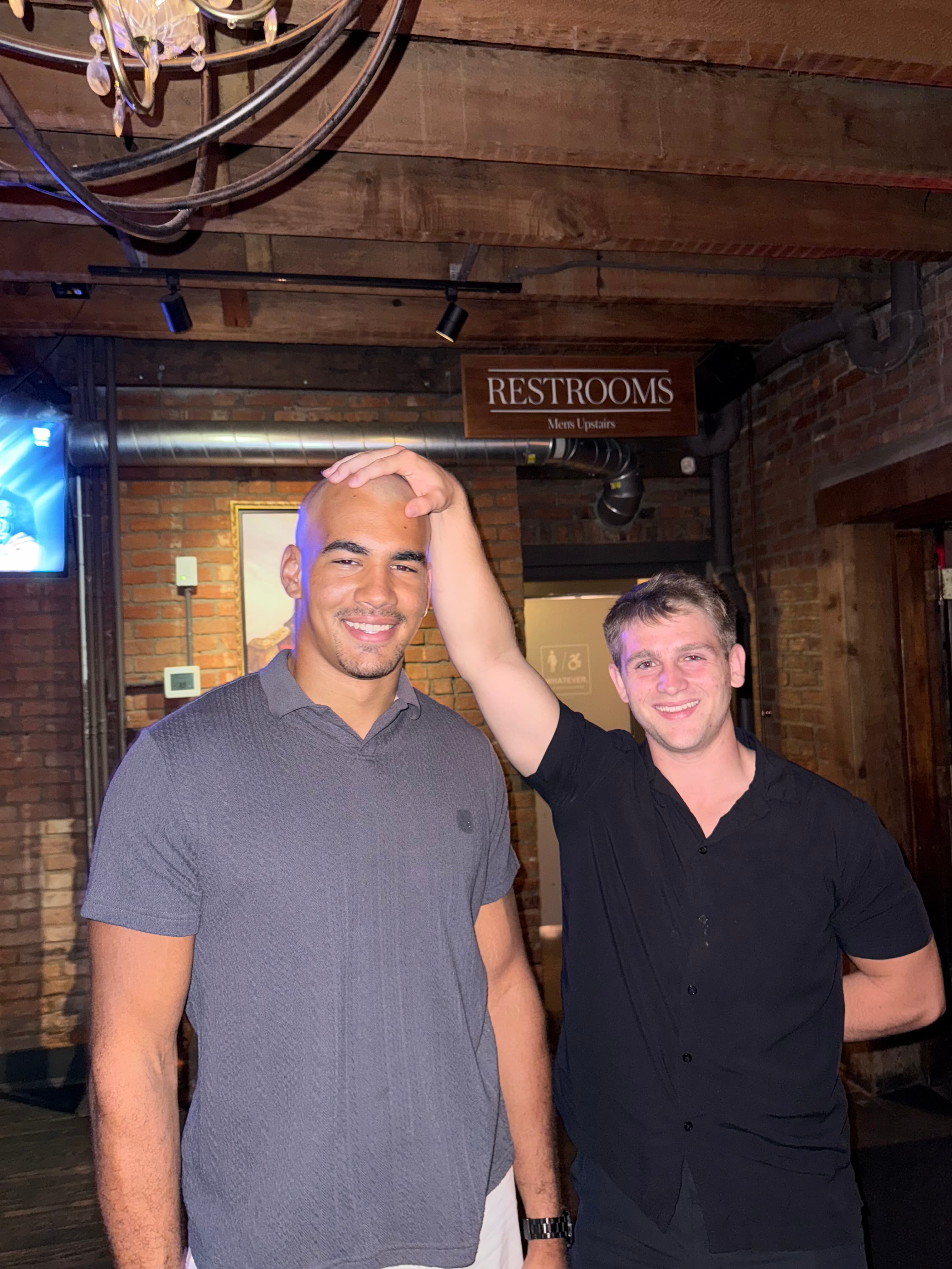 Two men smile as one playfully places his hand on the other's bald head.